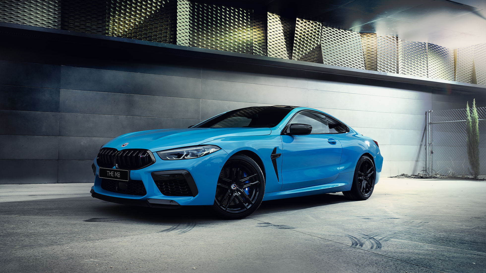 BMW M8 Competition Coupé, three-quarter front view. BMW M8 Competition Coupé in Marina Bay Blue metallic with shining LED headlights, standing three-quarter front view in front of villa at night.